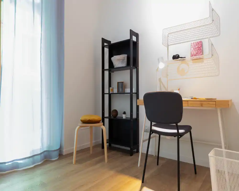 Rincón luminoso y minimalista de salón/trabajo con escritorio, silla y estantería abierta junto a una ventana con visillo — un espacio funcional y sereno.