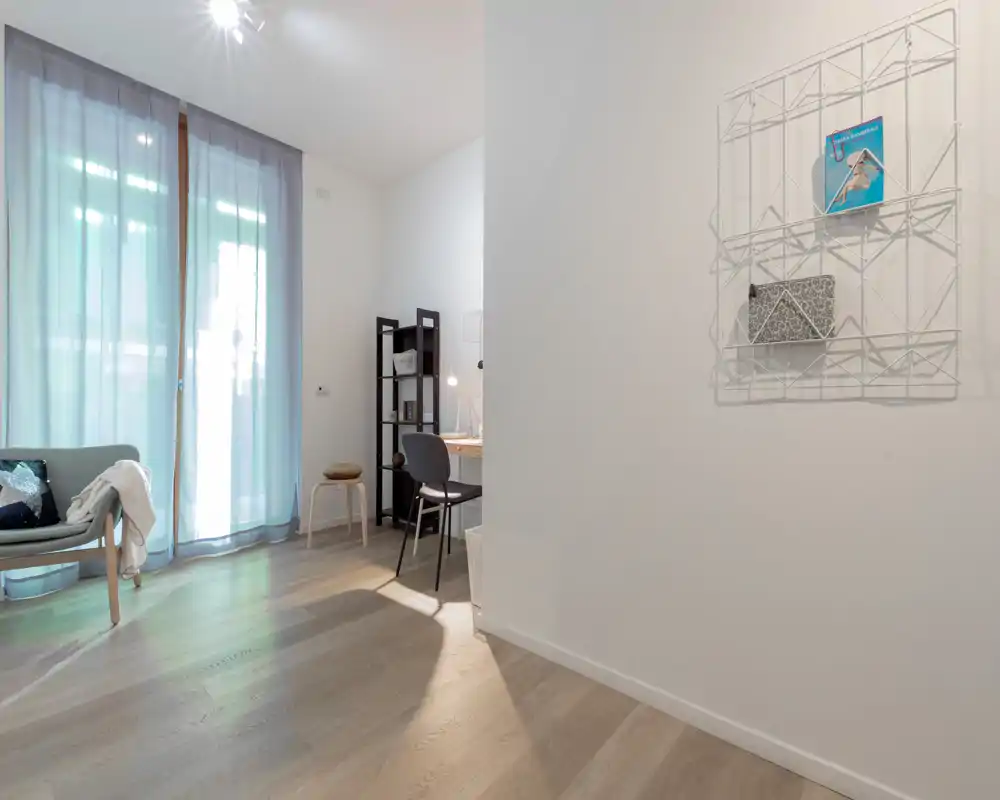 Área de estar moderna y luminosa con un sofá pequeño junto a una gran ventana y un rincón de escritorio compacto; estilo minimalista y luz natural que crean un ambiente acogedor.