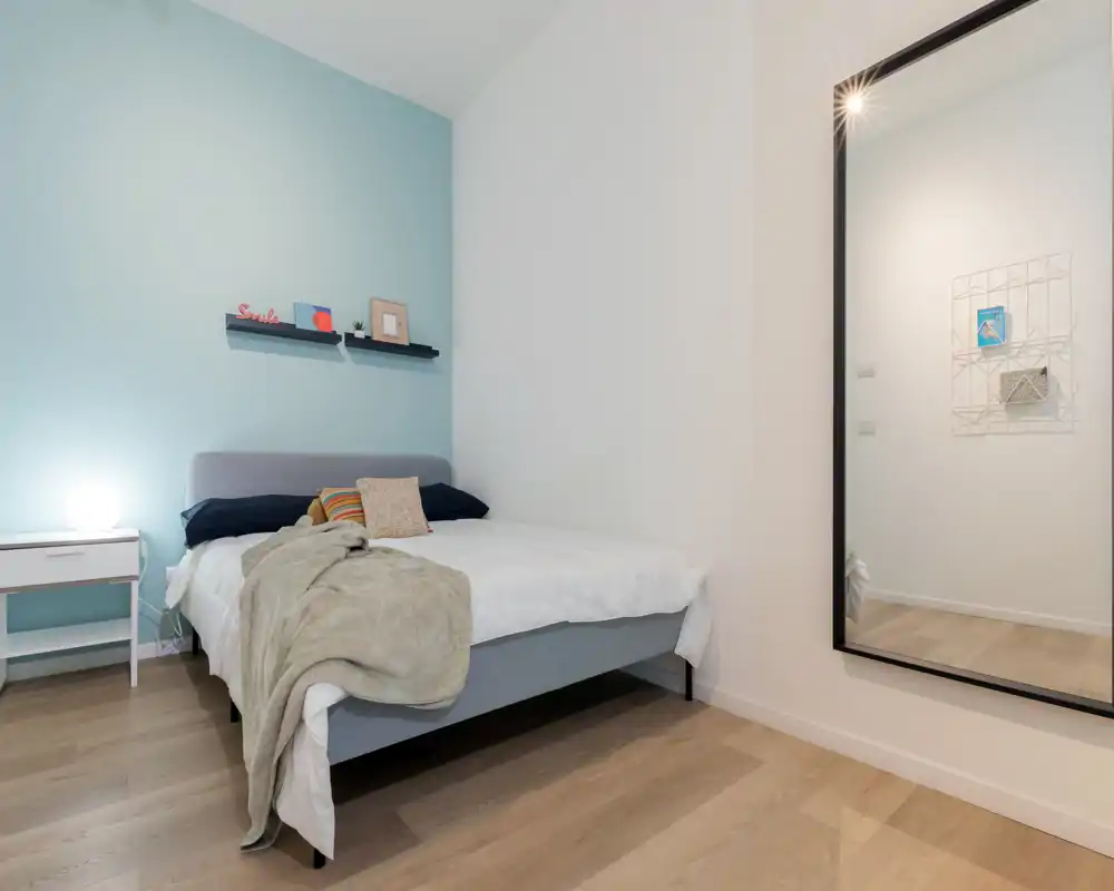 Dormitorio moderno y luminoso con cama doble bien hecha, manta y cojines, mesita con lámpara y un gran espejo en la pared que aporta profundidad.