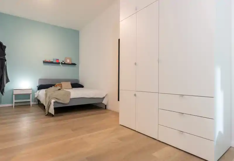 Dormitorio minimalista con cama doble frente a una pared de acento turquesa claro, mesita de noche con lámpara y un gran armario blanco empotrado; suelos de madera y textiles suaves crean una atmósfera moderna y tranquila.