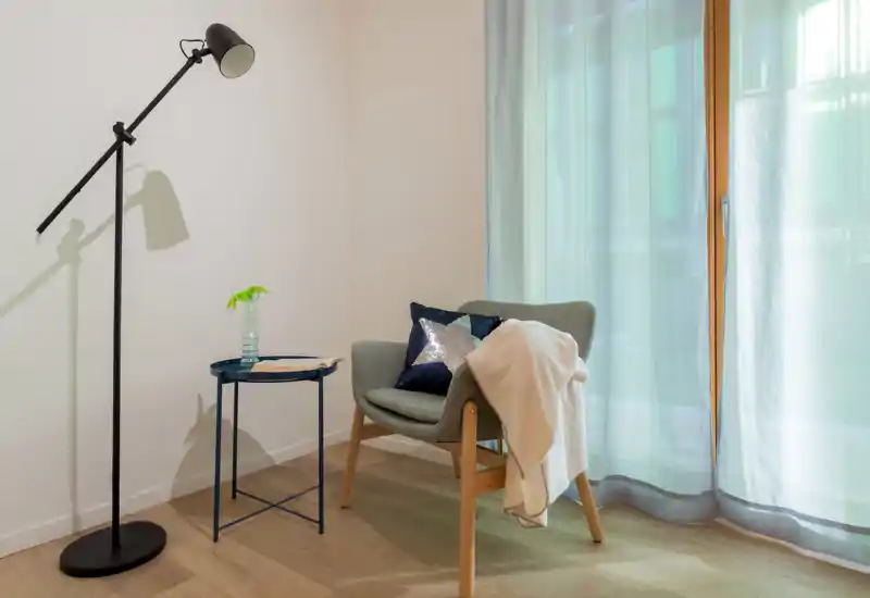 A cozy reading corner with a modern armchair, floor lamp and side table next to sheer-curtained windows — bright, calm and well styled for a relaxing stay.