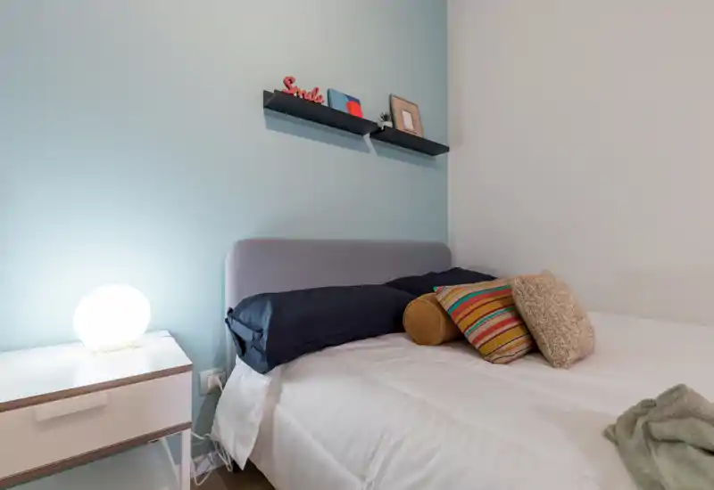 Bright, modern bedroom corner with a neatly made bed, decorative cushions and a bedside table with a soft-glow lamp — calm pastel wall and floating shelves add a cozy, styled touch.