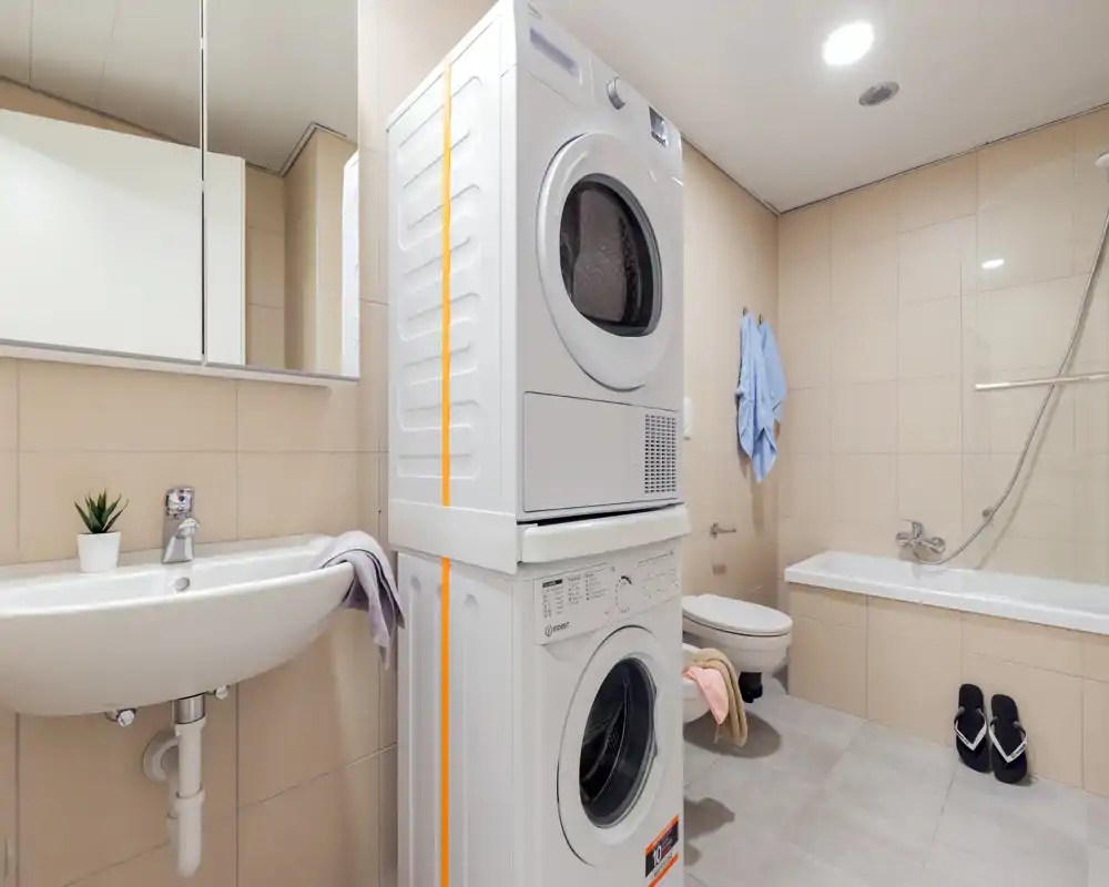 Baño moderno y luminoso con bañera, lavabo suspendido, inodoro y una lavadora/secadora apiladas — uso eficiente del espacio en un apartamento compacto.