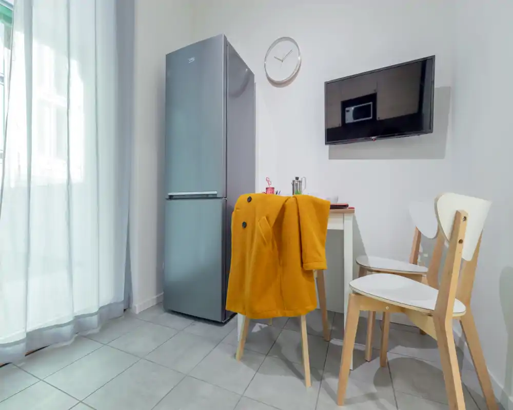 Rincón de cocina compacto con nevera alta de acero, mesa pequeña con sillas y televisor de pared; luz natural brillante y mobiliario moderno y minimalista.