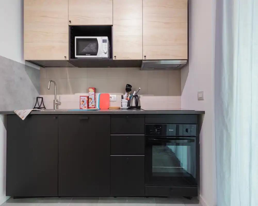 Kitchenette moderna y compacta con muebles bajos negros, armarios altos de madera, horno y microondas integrados; encimera ordenada con hervidor y accesorios — espacio luminoso y funcional ideal para alquileres de corta estancia.