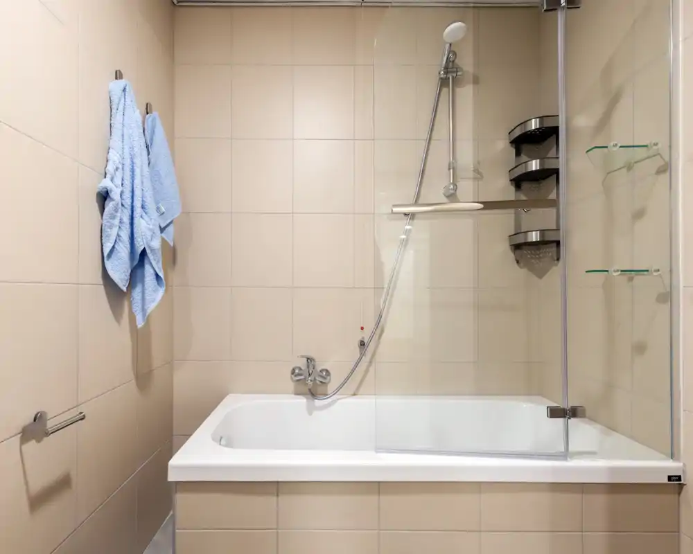Baño moderno alicatado con bañera empotrada y ducha de mano, mampara de vidrio, estantes de pared y dos toallas azules colgadas.