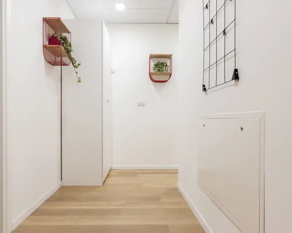 Entrada minimalista y bien iluminada con suelo de madera clara, paredes blancas y estantes sencillos; primera impresión ordenada y moderna.
