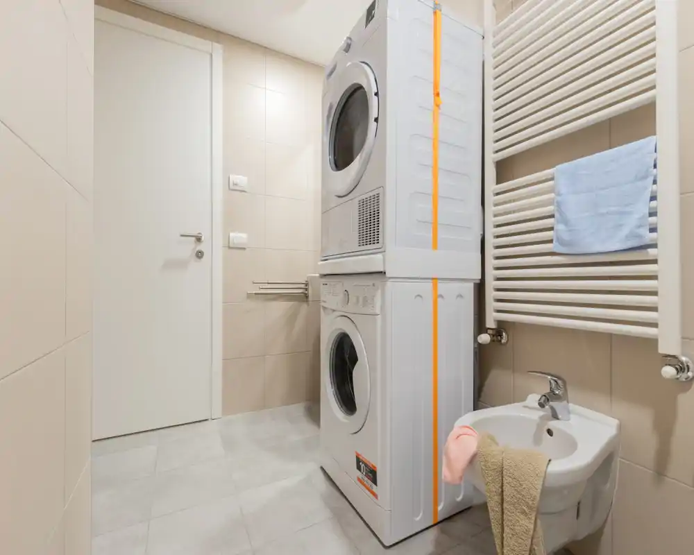 Baño compacto con lavadora y secadora apiladas, radiador toallero de pared y bidé; azulejos neutros e iluminación brillante que aportan un aspecto limpio y funcional.