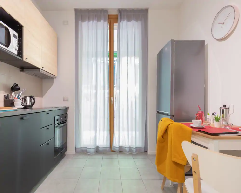 Cocina moderna y compacta con horno integrado, frigorífico y un rincón comedor pequeño. Las cortinas dejan pasar luz natural suave y los acentos cálidos hacen el espacio acogedor.