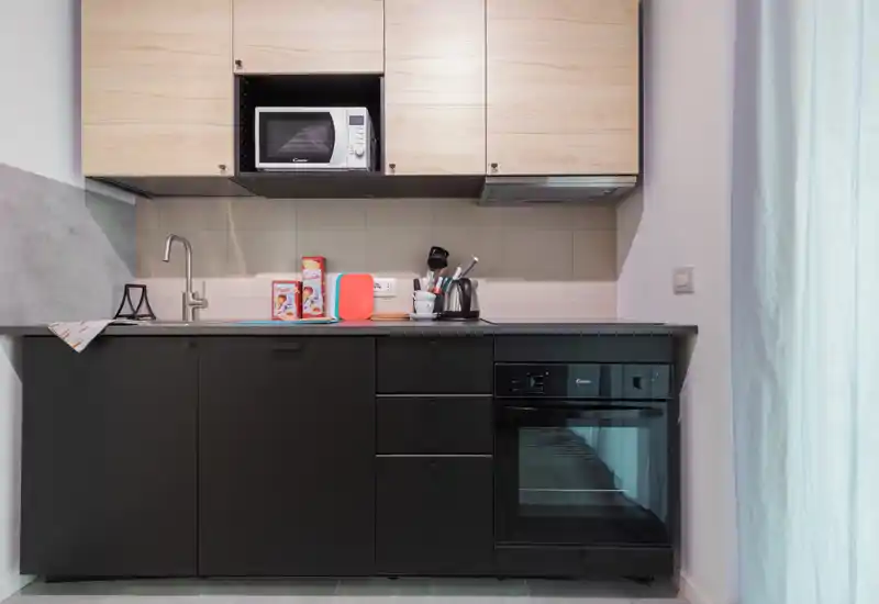 Compact modern kitchenette with black lower cabinets, wooden upper cupboards, built-in oven and microwave; tidy countertop with kettle and accessories — bright, functional space ideal for short-stay rentals.