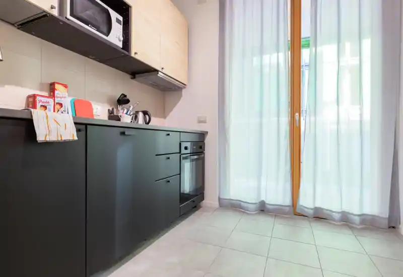 Modern compact kitchen with dark lower cabinets, built-in oven and microwave, kettle and utensils on the counter. Natural light enters through a large glass door with sheer curtains, creating a bright, inviting space suitable for a small apartment.