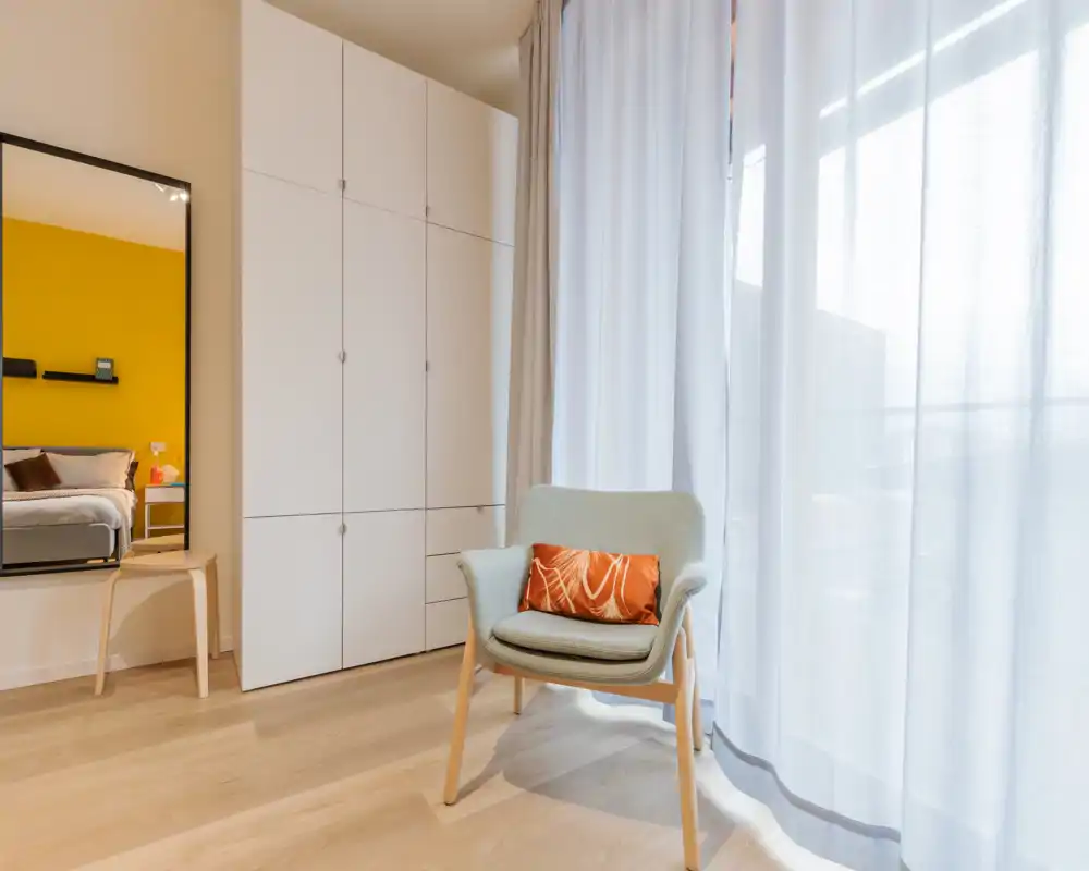 Bright, modern bedroom corner with a comfortable accent chair by a large sheer-curtained window; a mirror at left shows the bed and a warm yellow accent wall.