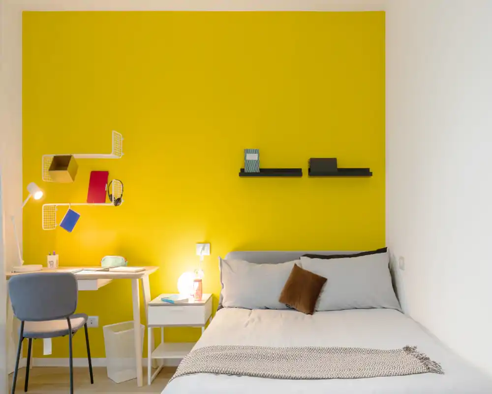 Bright, modern bedroom with a bold yellow accent wall, comfortable double bed, bedside table with lamp and a small work desk — ideal for a cozy stay.