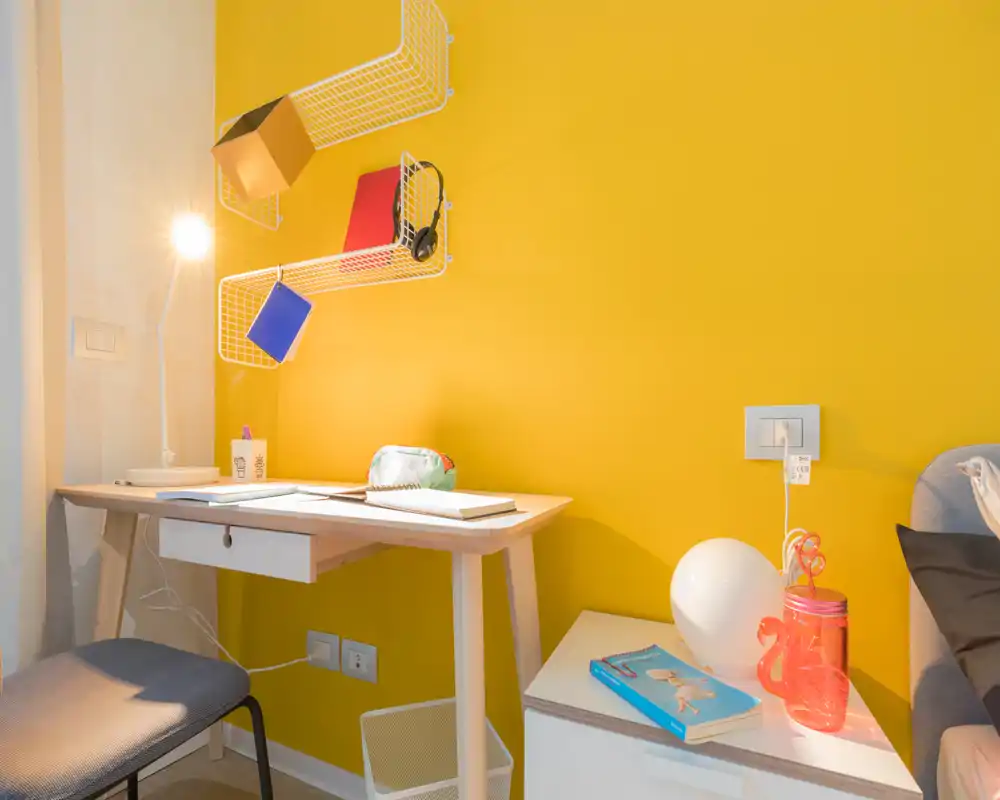 Small bedroom corner with a study desk and bedside table against a bright yellow wall, bedside lamp and decorative items create a lively, functional space.