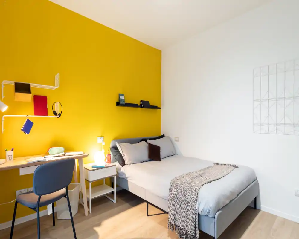 Bright, modern bedroom with a grey upholstered double bed, cozy throw and pillows, a bedside table and a small desk against a vivid yellow accent wall — clean, well-lit and styled for comfort.