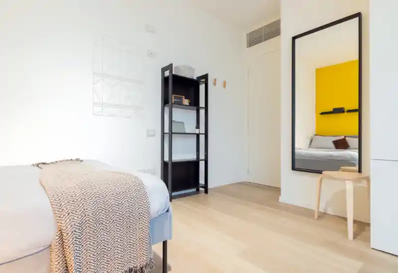 Dormitorio moderno y luminoso con cama bien hecha, manta texturizada y un espejo alto que refleja una pared de acento amarilla. Suelo de madera claro y estantería minimalista que crean un ambiente acogedor y despejado, perfecto para alquileres temporales.