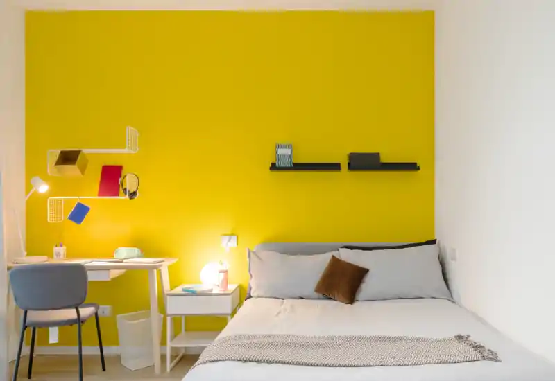 Dormitorio moderno y luminoso con una pared amarilla intensa, cama doble cómoda, mesita con lámpara y un pequeño escritorio — perfecto para una estancia acogedora.