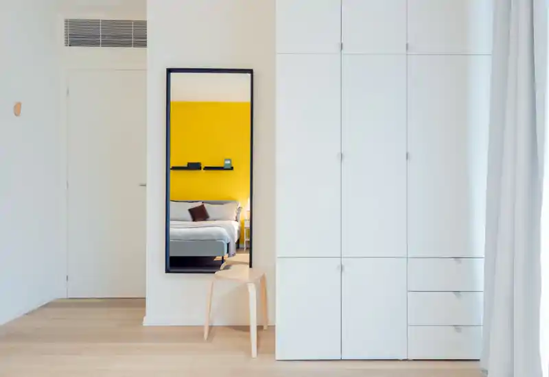 Dormitorio moderno y luminoso con un gran armario blanco y un espejo que refleja la cama frente a una pared amarilla. Estilo minimalista y luz natural que aportan una sensación fresca y acogedora.