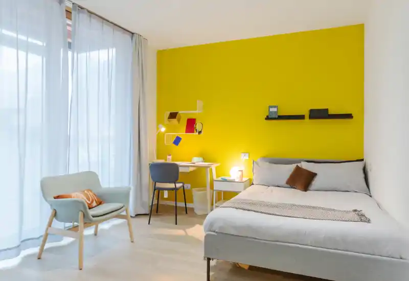 Dormitorio moderno y luminoso con cama grande, mesita y escritorio frente a una pared amarilla vibrante; cortinas translúcidas dejan pasar mucha luz natural y un sillón aporta un rincón acogedor.