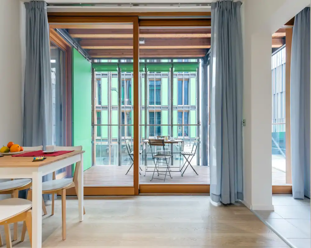 Bright modern dining area opening onto a covered balcony with a small outdoor table set. Clean wooden floors, simple table and chairs, and large sliding glass doors provide lots of natural light.
