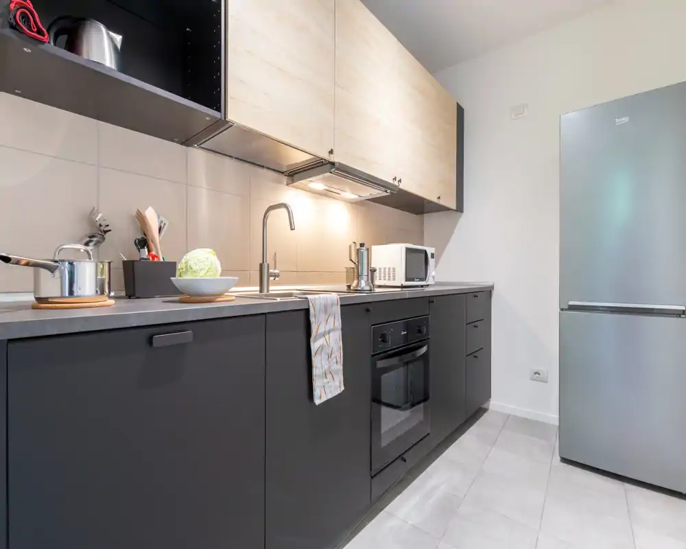Modern fitted kitchen with dark lower cabinets and light wood upper cabinets, equipped with an oven, induction hob, microwave and refrigerator. Clean, minimal styling with practical worktop and soft under-cabinet lighting.