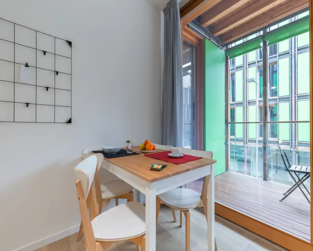 Small bright dining area with a wooden table for two, modern chairs and placemats, opening onto a glass-door balcony that brings abundant natural light.