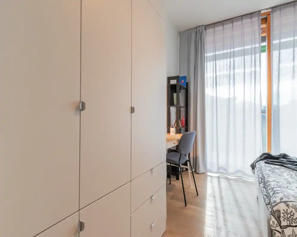 Bright, modern bedroom with large wardrobe, a study desk and a partially visible bed by a floor-to-ceiling window letting in soft natural light.