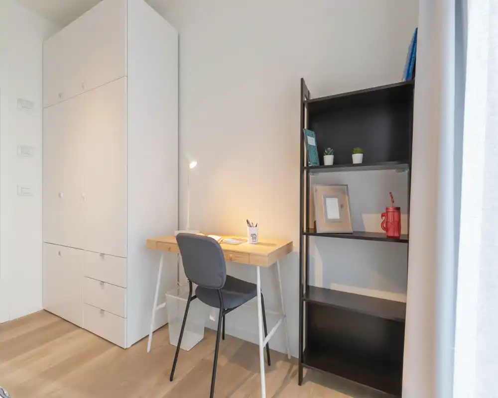 Bright, minimal bedroom corner with a wooden desk, chair and tall wardrobe; shelving with small plants and decorative objects adds a tidy, modern touch.