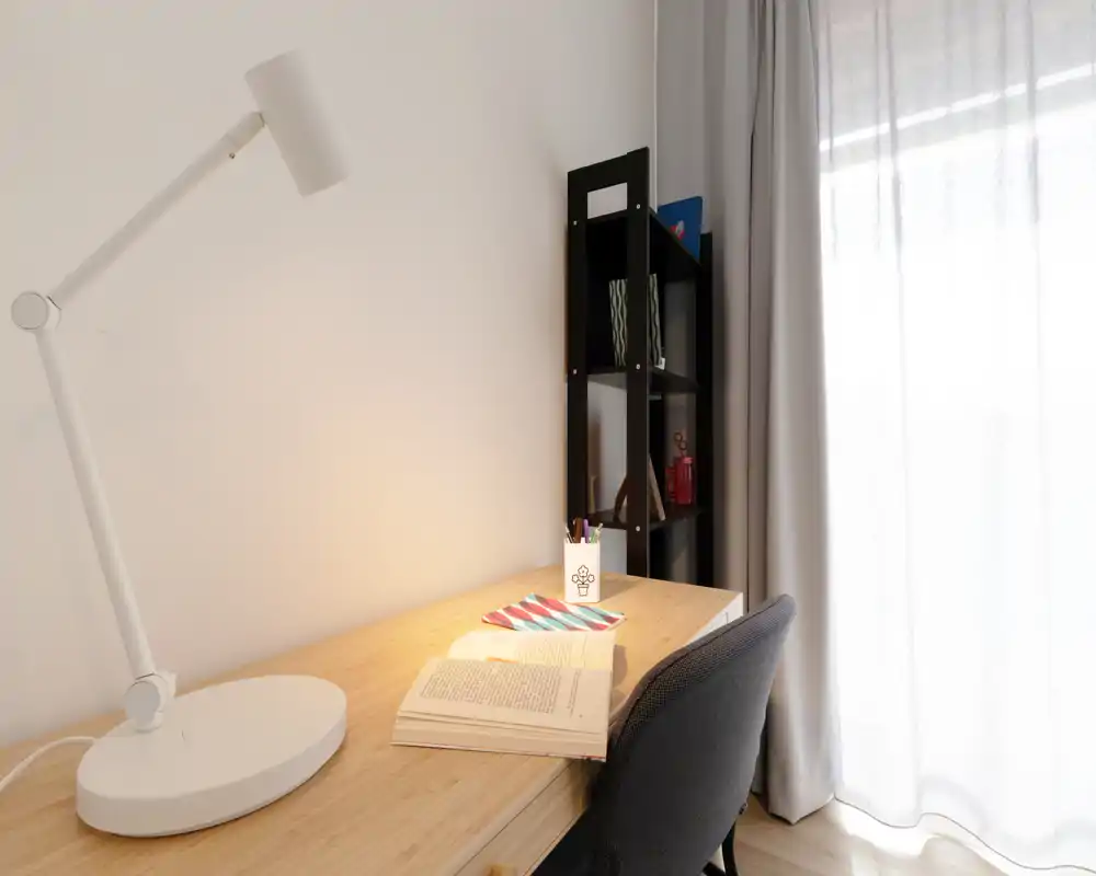 Bright, minimal desk/workspace with a lamp, chair, open book and shelving beside a large sheer-curtained window — ideal for a quiet home office or study corner.