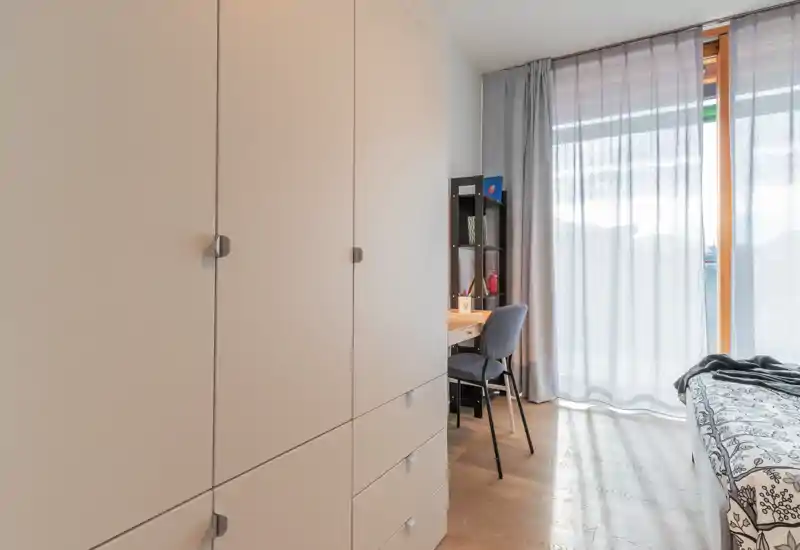 Bright, modern bedroom with large wardrobe, a study desk and a partially visible bed by a floor-to-ceiling window letting in soft natural light.
