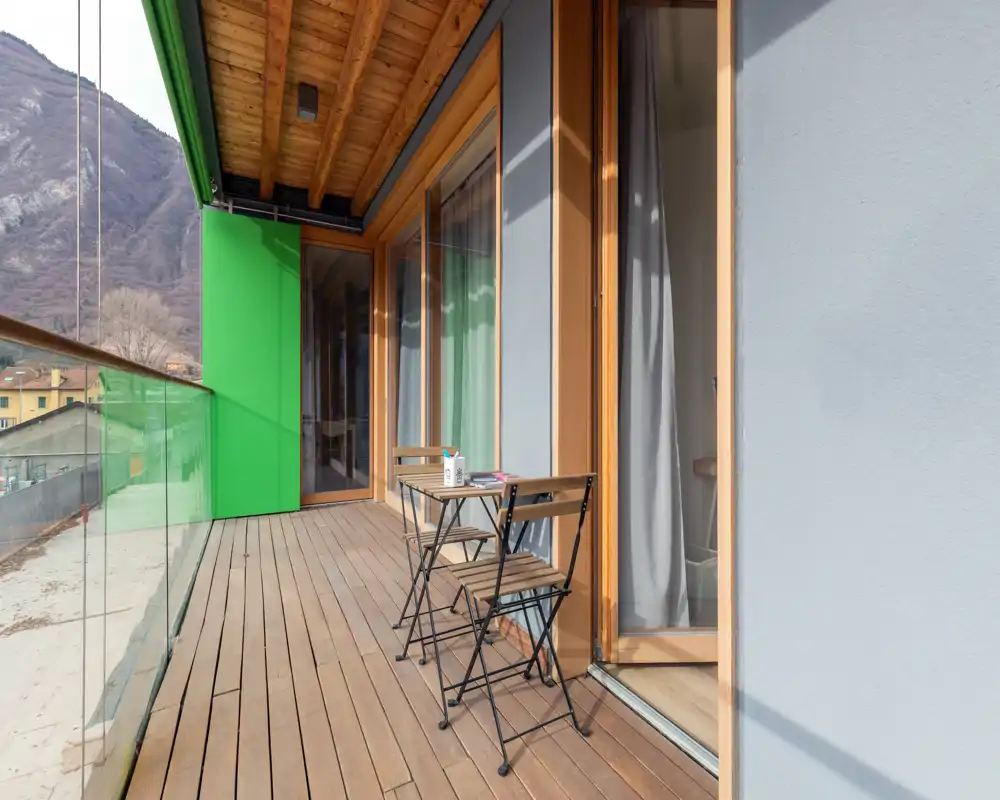 Sunny balcony with wooden decking, glass railing and a small bistro table with two chairs; overlooks a mountain view and connects to the interior through large sliding glass doors.