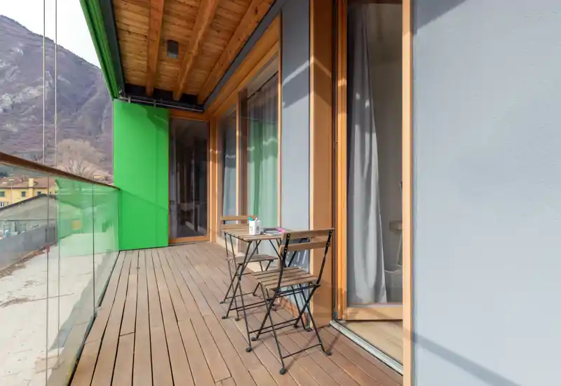 Sunny balcony with wooden decking, glass railing and a small bistro table with two chairs; overlooks a mountain view and connects to the interior through large sliding glass doors.