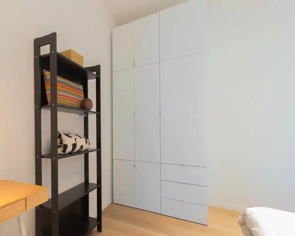 Angolo della camera piccolo e ordinato con armadio a incasso e bordo del letto visibile; pavimento in legno e mensole aperte per riporre oggetti.