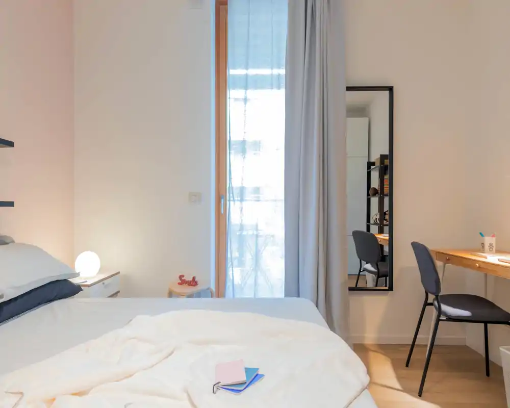 Camera da letto moderna e luminosa con letto ben rifatto, tessili morbidi e una piccola lampada sul comodino. Una scrivania e una sedia sono vicine a uno specchio intero e una tenda leggera lascia filtrare la luce naturale, creando un'atmosfera accogliente.