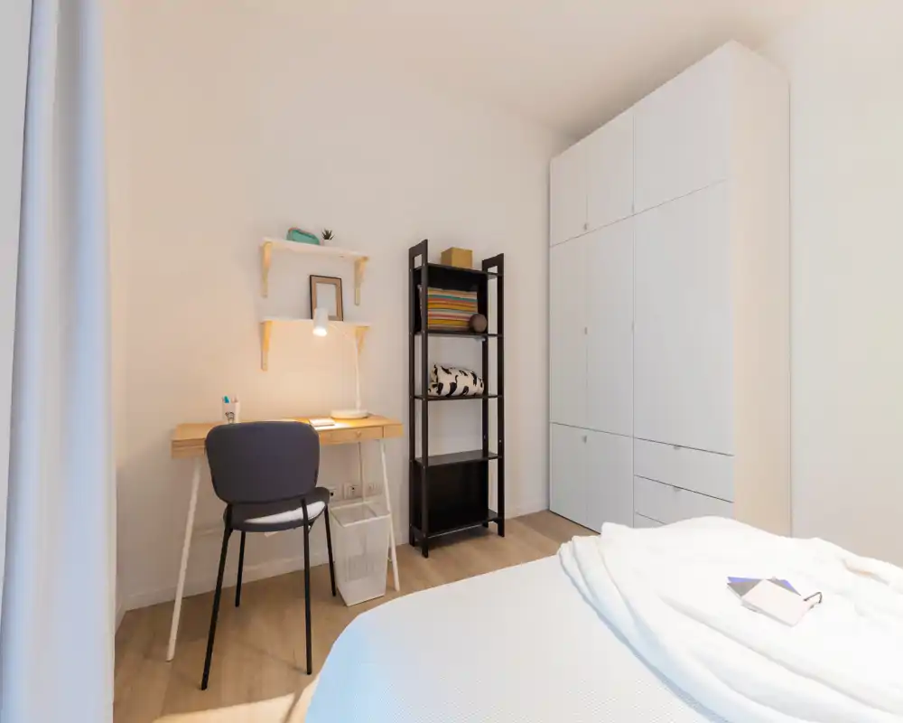 Camera da letto luminosa e minimal con letto in primo piano, una scrivania con lampada e sedia, mensole e un armadio bianco alto — ambiente pulito, moderno e accogliente.