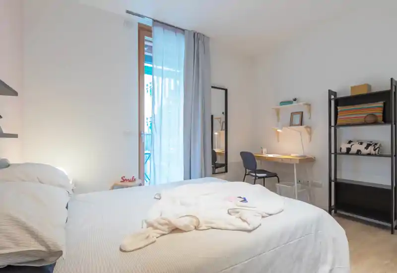Camera da letto luminosa e ben allestita con un grande letto, biancheria morbida, una scrivania e scaffali — ideale per un soggiorno confortevole con luce naturale dal balcone.
