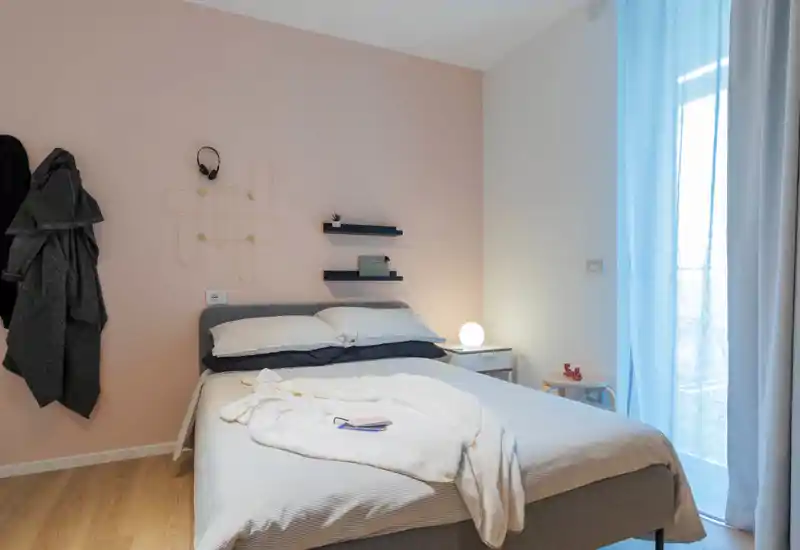 Camera da letto luminosa e accogliente con letto matrimoniale, biancheria morbida e lampada da comodino; parete pastello e luce naturale dalla finestra con tenda creano un'atmosfera tranquilla.