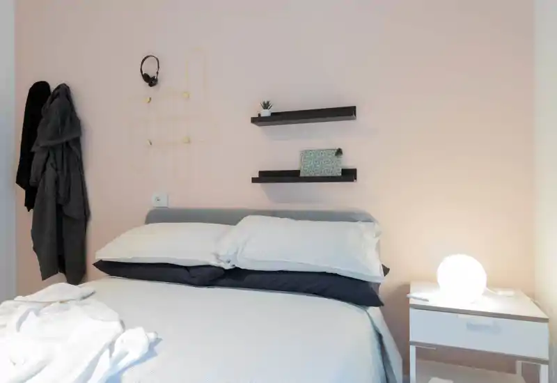Camera da letto moderna e luminosa con letto ben rifatto, cuscini morbidi e una lampada da comodino accesa; mensole minimali e tessuti accoglienti creano un'atmosfera tranquilla.