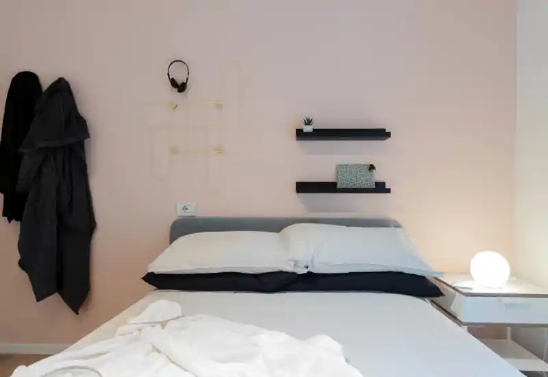 Camera da letto moderna e luminosa con letto matrimoniale ben rifatto, cuscini soffici e una lampada da comodino dal calore accogliente. Mensole minimal e appendiabiti aggiungono funzionalità e stile.