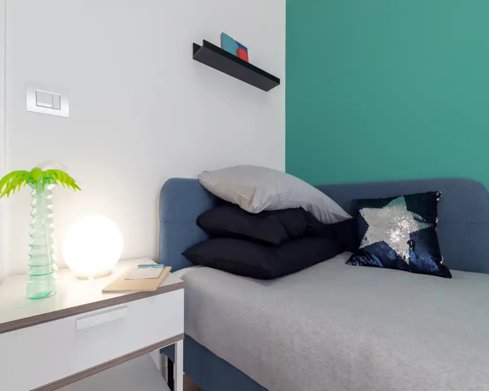 Angolo di camera luminoso e moderno con letto ben rifatto, cuscini decorativi e comodino con lampada e pianta — parete accentata verde acqua e mensola minimal.