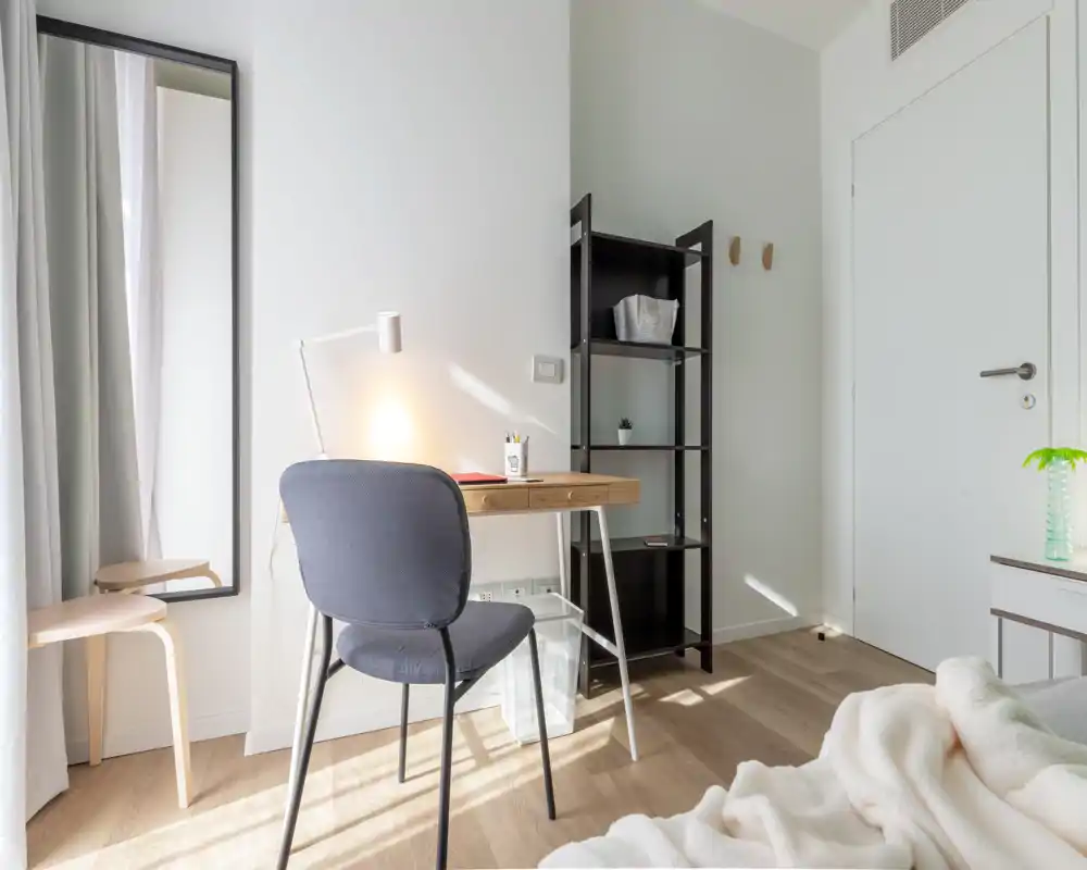 Angolo camera luminoso e minimal con una piccola scrivania, sedia e letto parzialmente visibile con biancheria morbida. Perfetto per un alloggio moderno e compatto con area lavoro e luce naturale.