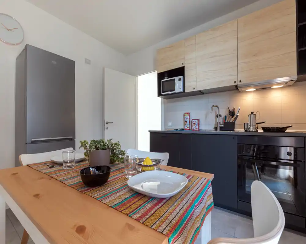 Cucina moderna ben attrezzata con forno incassato, microonde e frigorifero. Un tavolo in legno con tovaglia colorata e posti apparecchiati crea un'area pranzo accogliente.