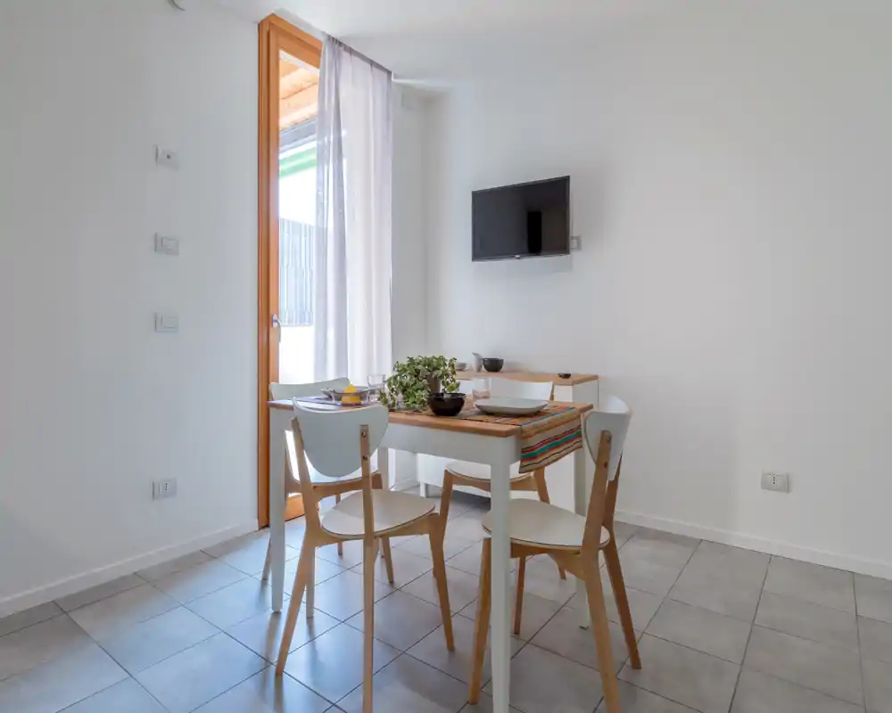 Zona pranzo luminosa e minimal con un piccolo tavolo in legno per quattro persone, runner decorativo e una pianta; la luce naturale dalla porta crea un'atmosfera accogliente.