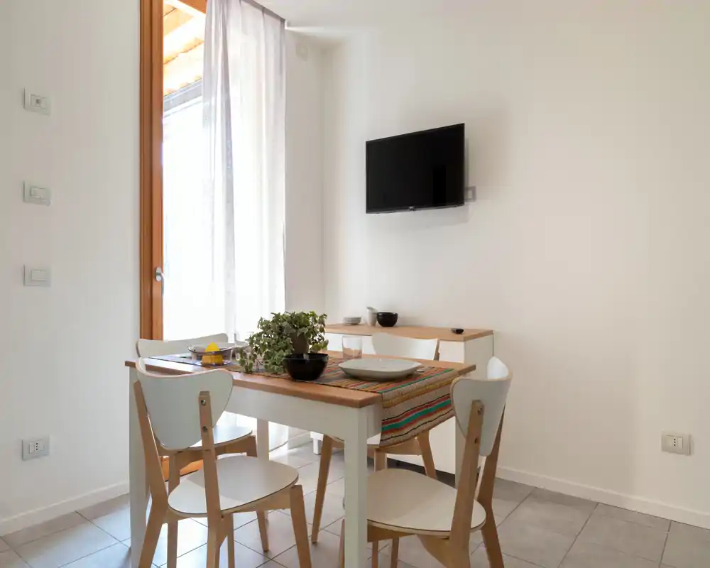 Zona pranzo luminosa e minimal con tavolo in legno per quattro, runner colorato, pianta decorativa e TV a muro — perfetta per pasti informali.