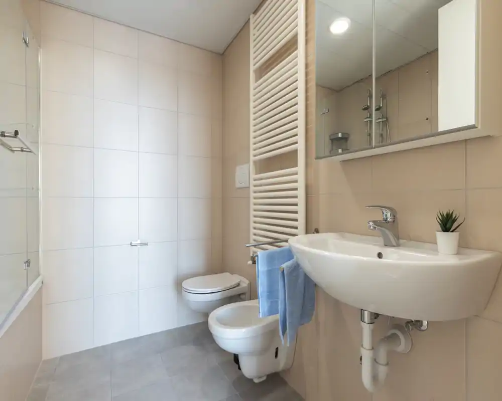 Bagno moderno e luminoso con wc e bidet sospesi, lavabo a parete con specchiera, scaldasalviette e piastrelle dai toni neutri per uno spazio pulito e funzionale.