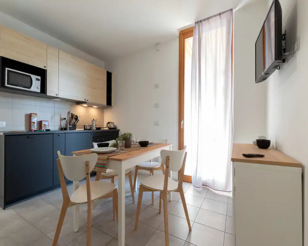 Cucina moderna a vista con tavolo in legno per quattro persone, pensili integrati, microonde e luce naturale da una porta finestra — luminosa e funzionale per soggiorni brevi.