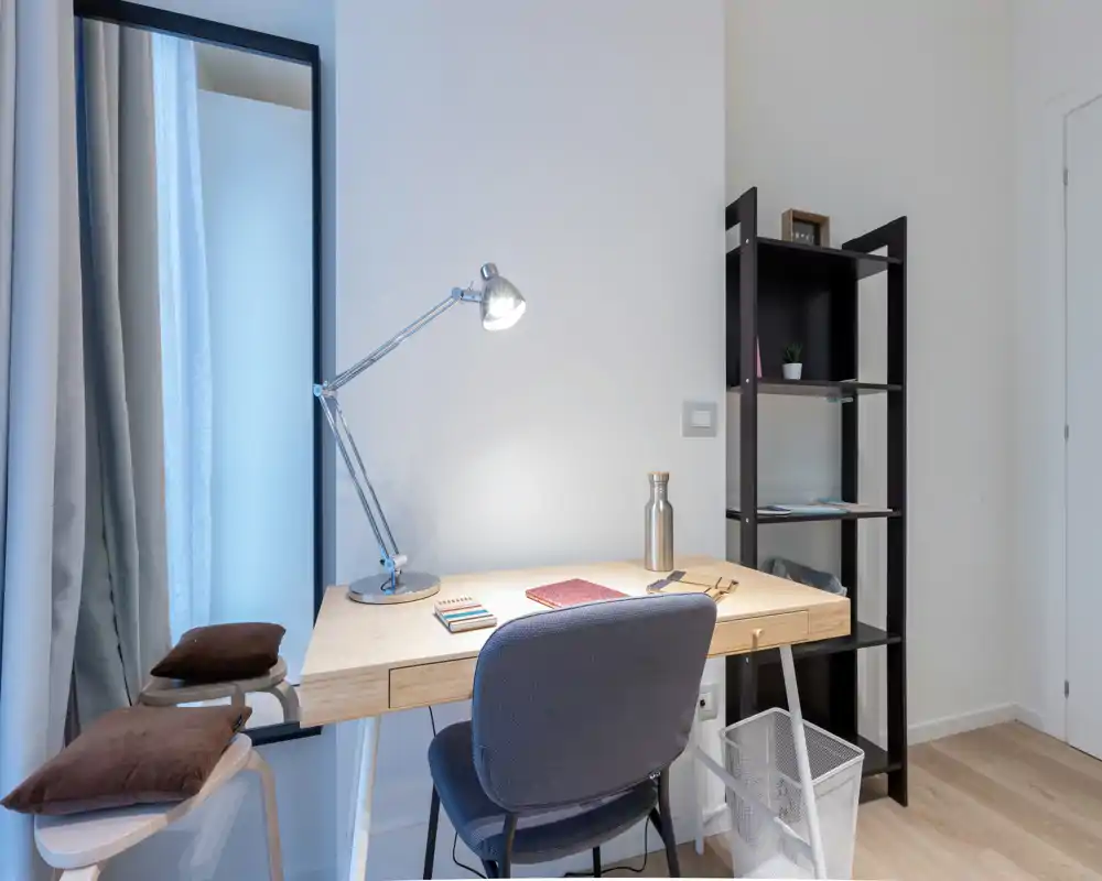 Rincón de trabajo compacto y luminoso en el salón: escritorio de madera con lámpara, silla acolchada, estantería y espejo que crean un espacio funcional y ordenado.
