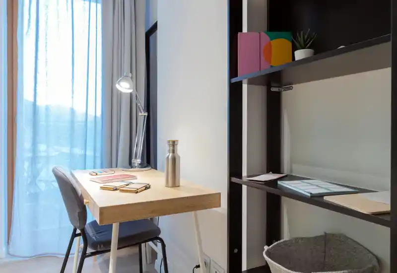 Agradable rincón de oficina en casa, luminoso y ordenado, con escritorio de madera, silla, lámpara y estanterías—perfecto para teletrabajo o estudio.