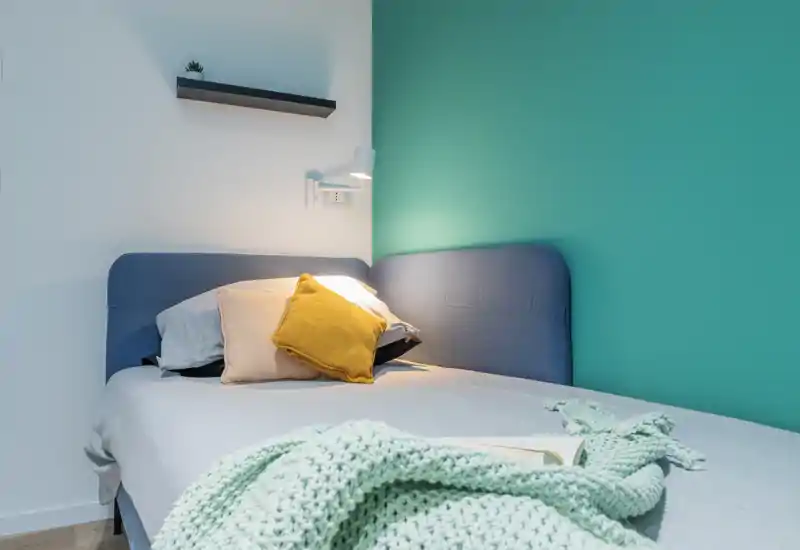 Dormitorio luminoso y acogedor con cabecero tapizado azul, cojines decorativos y una manta de punto. La luz tenue y la pared turquesa crean un ambiente fresco y agradable.