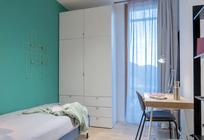 Dormitorio pequeño y luminoso con cama individual junto a una pared turquesa, un armario alto blanco y un escritorio junto a la ventana — ordenado, acogedor y ideal para un estudiante o una persona.